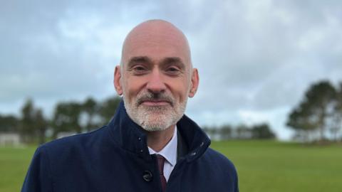 Professor Peter Bradley is wearing a navy overcoat with the collar turned up. He has a silver beard and is looking directly at the camera.