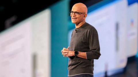 Satya Nadella smiling, wearing glasses and a grey sweater anbd standing in front of computer screens at a presentation