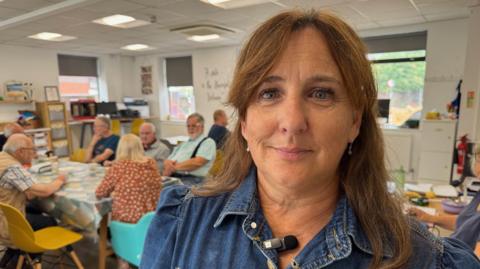 Isla looks at the camera, she has a fringe and brown hair down to her shoulders and is wearing a denim top. Behind her a mix of people can be sat around a dining table, the majority of them are elderly. 