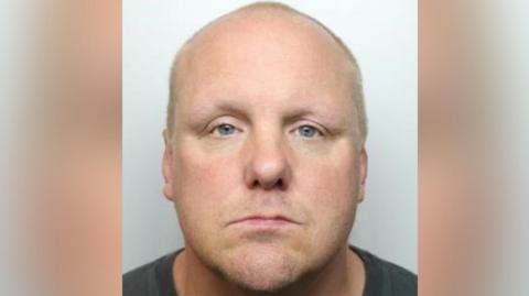 A mugshot of a man who looks directly at the camera. He has no facial hair and his hair is shaved.