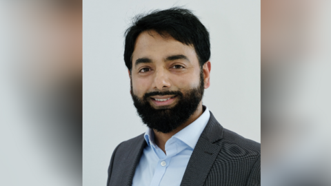 Ejaz Ahmed is stood in front of a grey background and is wearing a light blue buttoned shirt, with a grey jacket. He has short black hair and a black beard 