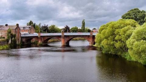 Three arches of a sandstone bridge over the River Nith are visible with trees to one side, cars crossing it and buildings to the left