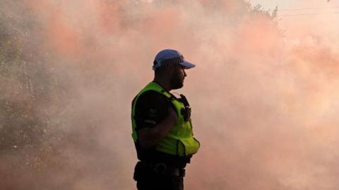 A police officer wearing a cap and hi-vis vest stands among a cloud of red smoke.