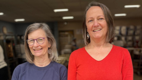 Jane Chong in a blue jumper, with glasses on and clear circular dangly earrings and Jane Kippax in a red jumper and silver round dangly earring. They both have hair to around their shoulders which is worn down and are smiling at the camera. The background is nondistinctive. 