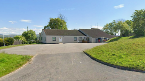 A grey building in Inverness with a black roof in the background with green grass to the right and left, in front of a bright blue sky.