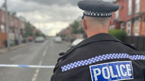 A police officer with his back to the camera wears a cap with a black and white square pattern on it and a black jacket with blue and white square edging, with the word "police" on it. He stands in front of police tape on a street