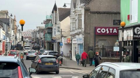 A picture taken from the end looking up at Camborne high street. There is a number of shops either side with people walking on pavements and cars driving up the street