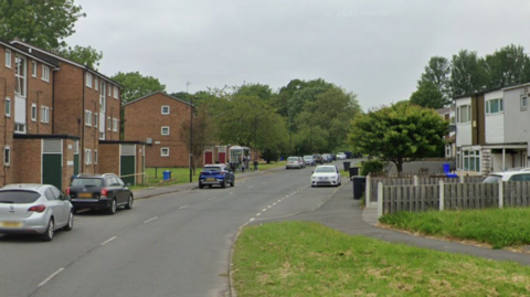A residential street. A few cars are parked around and short blocks of flats line the road.