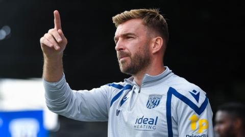 James Morrison holds is finger in the air from the sidelines during a game. He is wearing a West Bromwich Albon track top