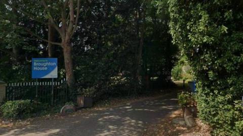 A Google Street View shot of the entrance to a care home with trees causing shadows on both sides of the path. A blue sign to the side of the path reads "Welcome to Broughton House".