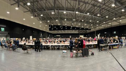 A wide view of a large hall which is being used for a vote count. There are rows of tables with people sitting at them