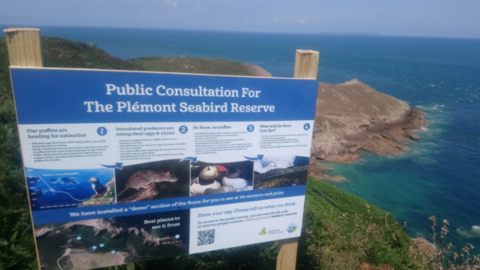 A sign with writing on it on a cliff with sea and a coastline behind.
