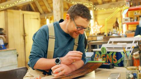 Alasdair wears an apron in a workshop with fairy lights. He is painting an oar on the table.