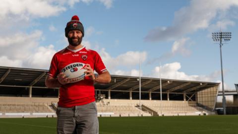 Salford city mayor Paul Dennett, stands on the pitch at Salford Community Stadium holding a rugby ball while wearing a red Salford Red Devils top and Salford Red Devils bobble hat with faded black jeans. 