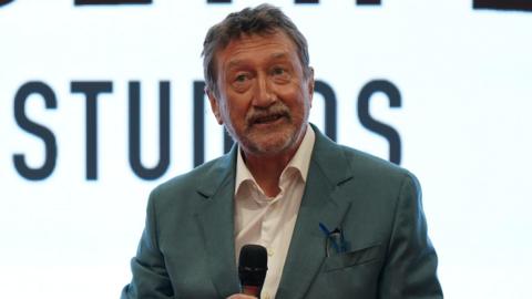 A man in a grey suit jacket and white shirt holds a microphone mid-speech. He is standing in front of a white backdrop with the word studios in capital letters. He has glasses tucked in his breast pocket, grey/brown hair and a neat greying beard.