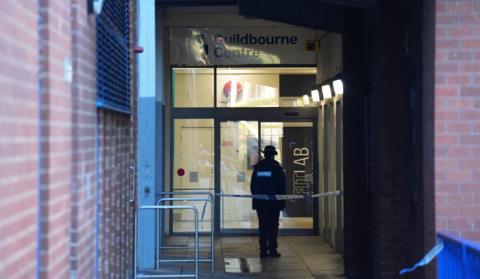 A lone police officer in uniform stands behind blue and white police tape outside a building