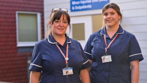 Two female nurses stand smiling outside a hospital building. 