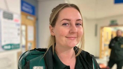 A close-up of a woman with blonde hair tied back, wearing a green parademic uniform