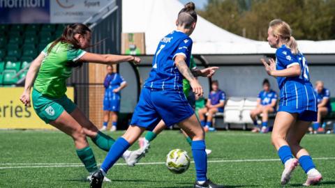 Calleigh Hedley (left) striking a shot with two Chessington & Hook United players in front of her. She is wearing a green strip and has tied back brown hair. The Chessington players are blue kits.