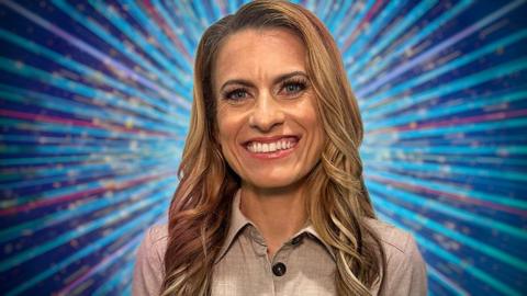 Karen has long honey blonde wavy hair. She is wearing a beige button-up top. She is smiling and standing in front of a blue background with purple, teal and pink stripes which fan out behind her, as well as gold glitter 