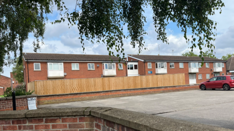 View of an estate made up of conjoined flats inside red bricked buildings. There is a wooden fence surrounding the back of the homes. One car is parked in the car park. On one of the walls is a piece of paper outlining the closure order terms.
