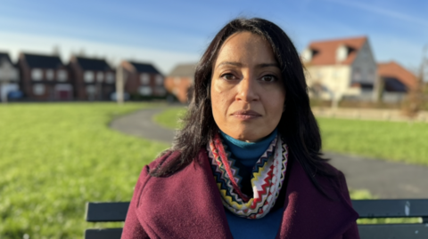 Khizra Bano sitting on a bench outside in an open space near some residential streets. She is wearing a maroon coat and blue polo neck with a brightly coloured scarf and has long black hair and is looking at the camera.