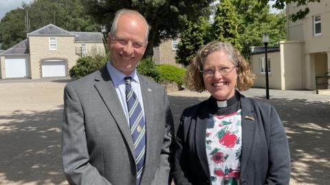 Jersey's Lieutenant Governor, Jerry Kyd, wearing a suit stands next to the Rev Sarah McClelland, a woman with glasses wearing a rector's dog collar.