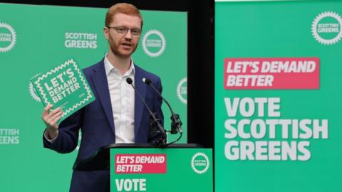 Ross Greer, who has short ginger hair, holds a green manifesto in his right hand while speaking into a podium. He is wearing a blue suit and white shirt, with green branding behind him