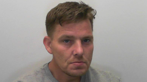 A mugshot of a man looking at the camera. He is wearing a grey top and has short brown hair. 