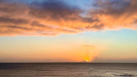 View of the sun rising over the sea. The sun, partially obscured by cloud, casts orange rays upwards into a sky tinged with peach and orange, over a darker grey sea.