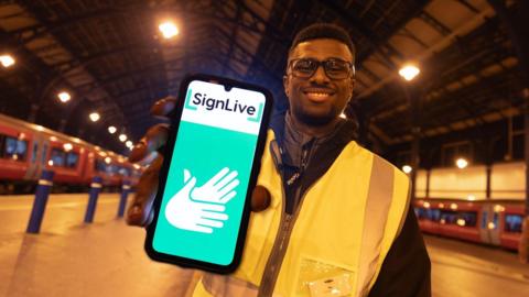 A person in a high vis jacket in a train station smiling and holding a phone with the SignLive logo on it.