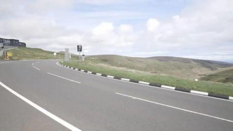 A close up of the curving Mountain Road with a building on the left and hills in the background.