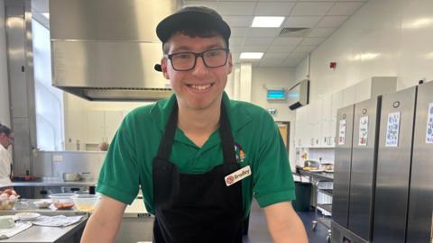 Bradley stands in the kitchen and wears a green T-shirt, black apron with his name badge on it and a cap that covers his short dark hair. He is wearing glasses and is smiling.