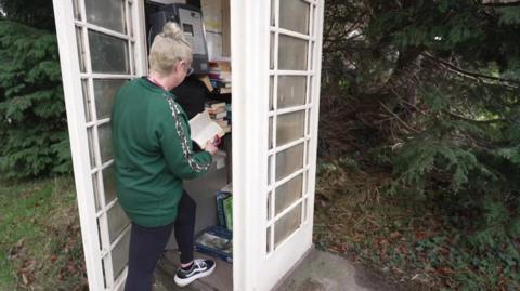 A woman reads a book as she stands in the doorway of a mid-20th century cream-coloured telephone box. More books can be seen stacked inside, alongside a pay phone. The woman has blond hair, tied in a bun, and wears a green top and black leggings.