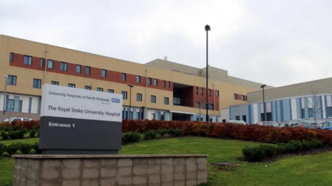 The front entrance of The Royal Stoke University Hospital. The beige and brown coloured building is in the background with a grassy mound in the foreground, with a sign on the left place on a brick wall.