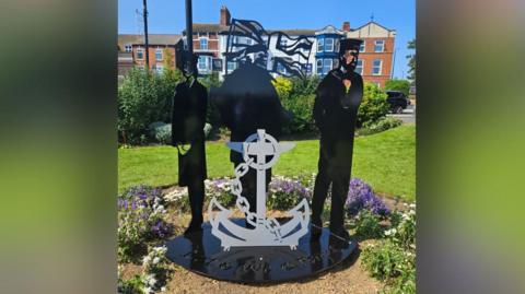 An image of the new memorial, it features a Navy wren, sailor and Royal Marine with an anchor in the middle. The memorial is made from black and grey sprayed metal cut outs and is placed in the middle of a flower bed.