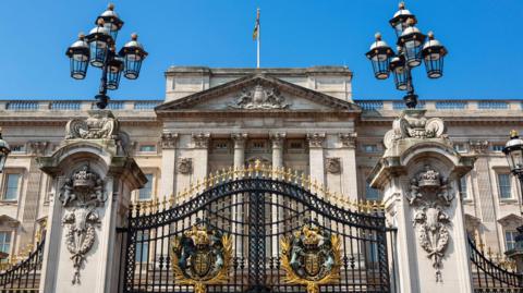 Exterior of Buckingham Palace. It is a large stone-coloured building with columns and a balustrade at the top. In the foreground, there are two large pillars with lanterns on top of them and ornate black and gold gates in the middle. 