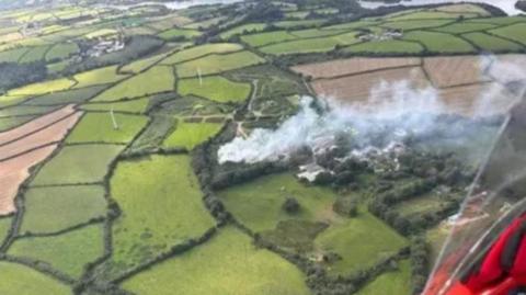 Aerial image of smoke belching upwards into the atmosphere from a compost fire on the ground, which is surrounded by green and verdant arable crop fields and in the distance, the gentle bend of a river or lake along with an enticing glimpse of a light plane fuselage painted in a rich and attractive red hue.