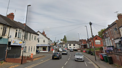 A road with several cars driving along it and buildings on either side of it. There is a black and white average speed check sign to the left.