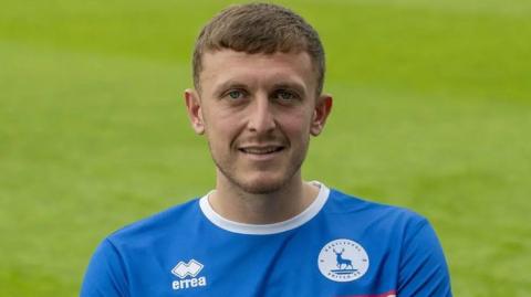 Football player Oliver Finney. He is wearing a blue Hartlepool United shirt and is stood on a football pitch.