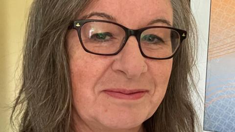 A close-up photo of a long haired woman with black glasses smiling at the camera.