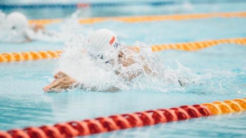 A swimmer with their arms outstretched and wearing a cap. The pool has red and yellow ropes marking the swimming lanes.