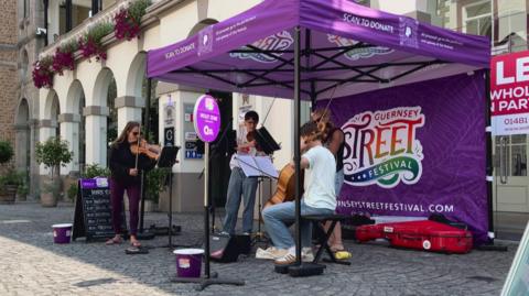 A group of musicians playing instruments under a purple gazebo on the street in front of some shops. There are collection buckets on the floor in front of them. 