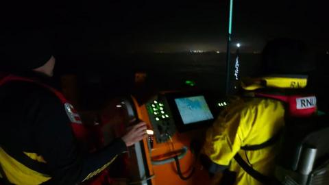 Two RNLI crew members check their radar as they looks for a missing boat at night. Both crew have yellow clothing and red lifejackets on. Lights from Jersey's coast can be seen in the distance.