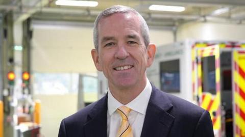 Neill Moloney looking towards the camera smiling. He has short grey hair and is wearing a black suit jacket, a white shirt and yellow tie. 