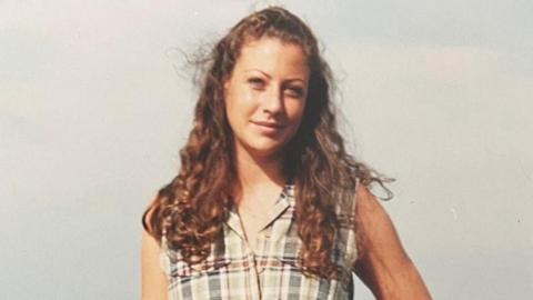 A photo of a young woman with long auburn curly hair wearing a pale checked sleeveless top 
