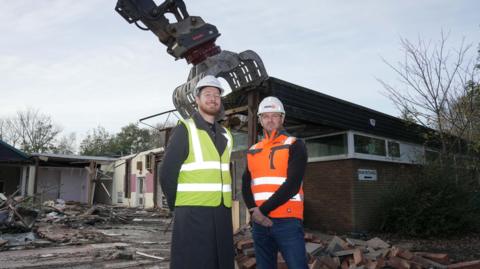 Two men standing in white hard hats and fluorescent jackets on a demolition site. A pile of bricks is behind them and a demolition machine is above a one-storey red brick building.