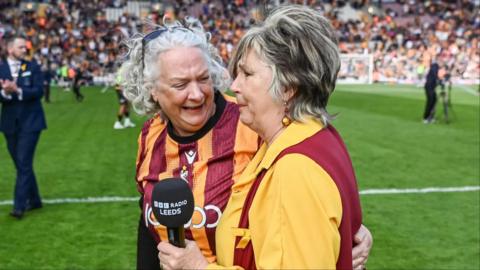Opera singer Lesley Garratt holds a BBC Radio Leeds branded microphone and has an emotional facial expression. A female member in a Bradford City football top of the Bantam of the Opera choir smiles and talks to Lesley as they stand on the football pitch.