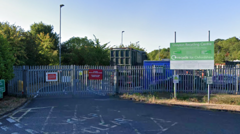 The metal gates outside a recycling centre. A container can be seen past the fence. A green sign outside reads "Drayton Recycling Centre".