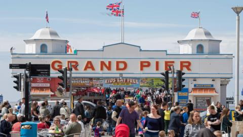 The Grand Pier in Weston-super-Mare. The pier has a white arch and union jack flags on top of it. "Grand pier" is written across the top in red capital letters and there are various signs advertising food and drink stalls. There is a large crowd in front of the pier.